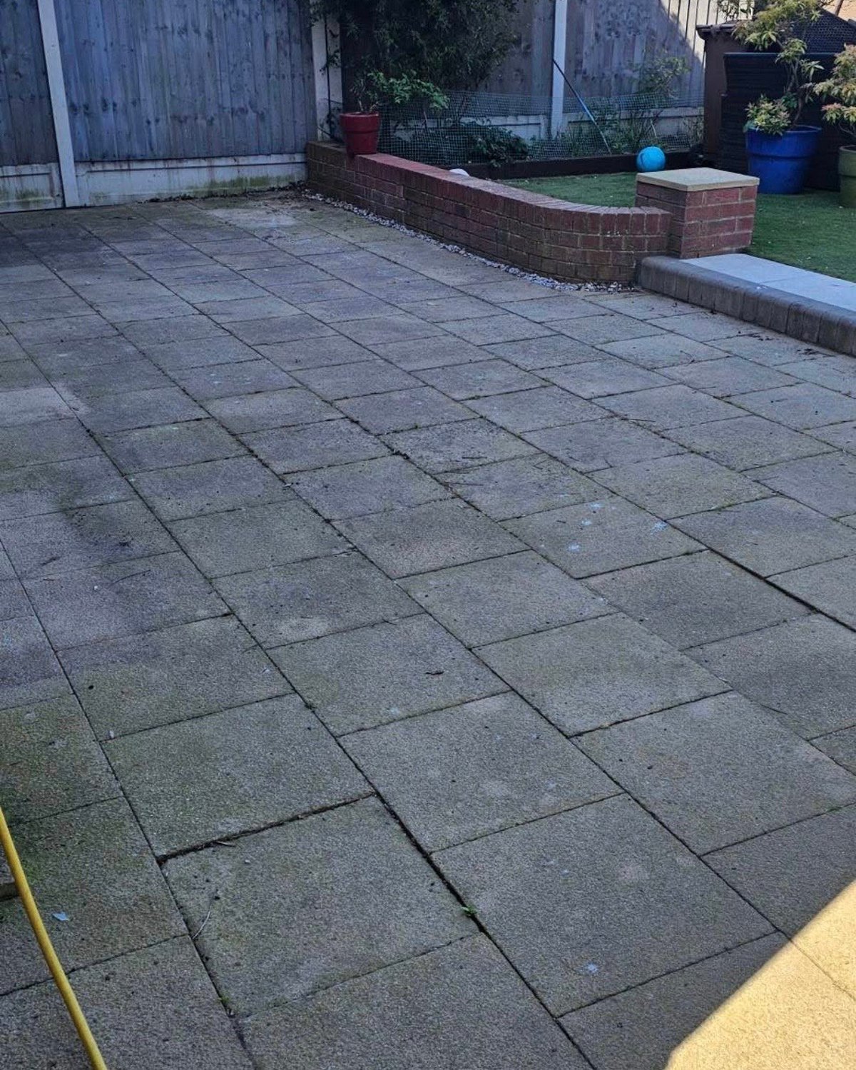 Gray paved patio with brick garden beds, metal fence, and blue bins in a residential backyard